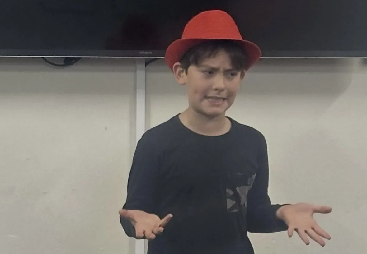 Student in a red hat presenting a script during a speech and drama class
