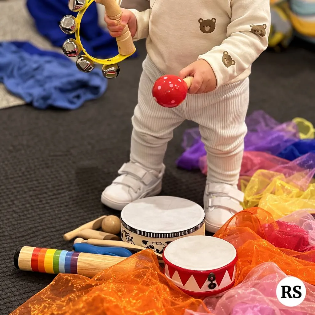 Toddler playing tambourine surrounded by colourful instruments and scarves in Tell and Show class
