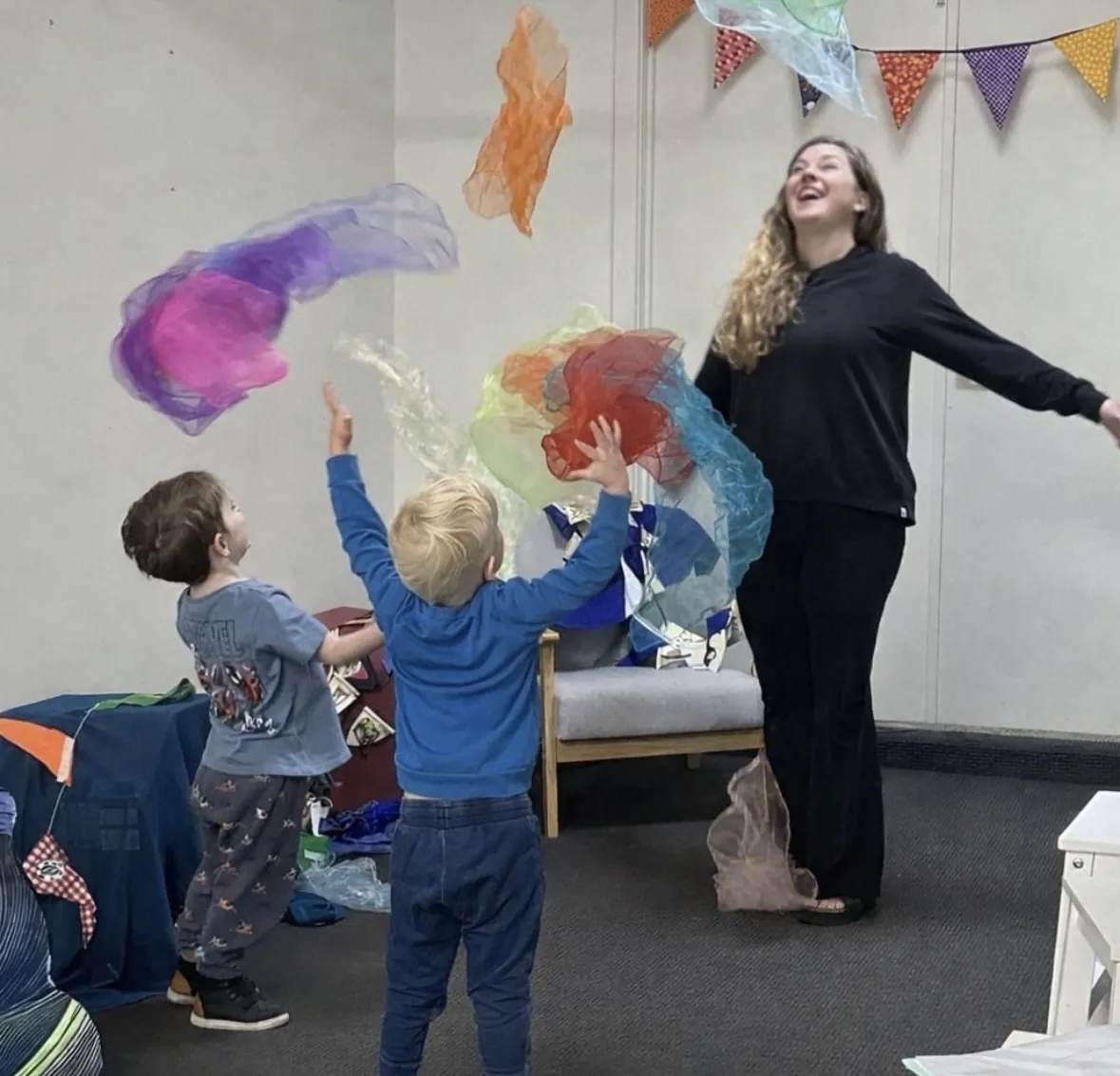 Young child and teacher playing with colourful scarves under bunting at a Tell & Show class